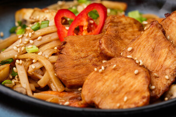 Udon noodles with teriyaki chicken and vegetables: zucchini, red pepper, mushrooms, carrot, onion and sesame seeds. Dish isolated in a blue bowl, close-up on a black marble background. Asian cuisine.