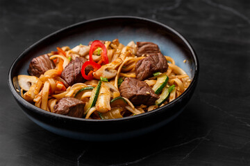 Udon noodles with teriyaki beef and vegetables: zucchini, red pepper, mushrooms, carrot, onion and sesame seeds. Dish isolated in a blue bowl, close-up on a black marble background. Asian cuisine.