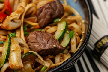Udon noodles with teriyaki beef and vegetables: zucchini, red pepper, mushrooms, carrot, onion and sesame seeds. Dish isolated in a blue bowl, close-up on a black marble background. Asian cuisine.