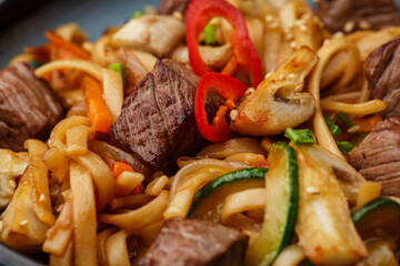 Udon noodles with teriyaki beef and vegetables: zucchini, red pepper, mushrooms, carrot, onion and sesame seeds. Dish isolated in a blue bowl, close-up on a black marble background. Asian cuisine.