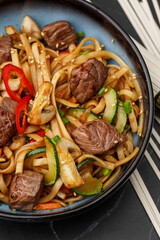 Udon noodles with teriyaki beef and vegetables: zucchini, red pepper, mushrooms, carrot, onion and sesame seeds. Dish isolated in a blue bowl, close-up on a black marble background. Asian cuisine.