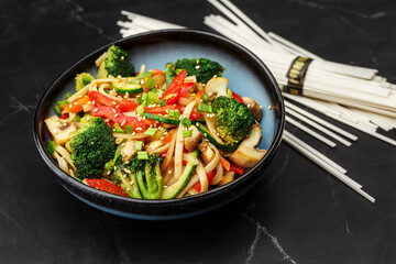 Udon noodles with vegetables: zucchini, broccoli, red bel pepper, mushrooms, carrot, spring onion and sesame seeds. Dish isolated in a blue bowl, close-up on a black marble background. Asian cuisine.