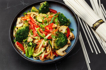 Udon noodles with vegetables: zucchini, broccoli, red bel pepper, mushrooms, carrot, spring onion and sesame seeds. Dish isolated in a blue bowl, close-up on a black marble background. Asian cuisine.