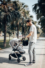 Young handsome father walking with his baby girl in the stroller around green park with palm trees