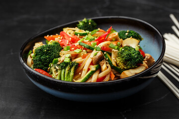 Udon noodles with vegetables: zucchini, broccoli, red bel pepper, mushrooms, carrot, spring onion and sesame seeds. Dish isolated in a blue bowl, close-up on a black marble background. Asian cuisine.