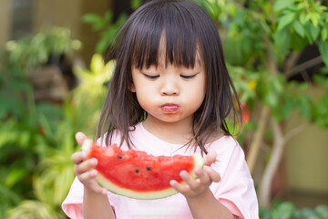 Portrait​ image​ of​ 2-3 yeas​ old​ of​ baby.​ Happy​ Asian child girl eating and biting a piece of watermelon. Enjoy eating moment. Healthy food and kid concept.​ Sucking fingers in the mouth.
