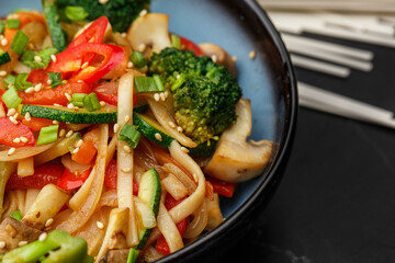Udon noodles with vegetables: zucchini, broccoli, red bel pepper, mushrooms, carrot, spring onion and sesame seeds. Dish isolated in a blue bowl, close-up on a black marble background. Asian cuisine.