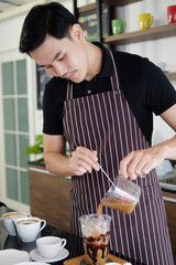 Smiling Asian barista young man pouring fresh milk in a plastic glass of latte iced coffee for according to order at counter bar in the morning at modern cafe.