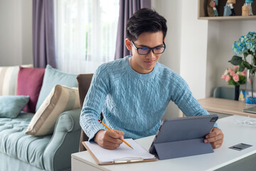 Young Asian man enjoy studying online using a tablet and taking notes at home.