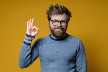 Cute, funny man in a sweater doing ok sign with hand and fingers, he is successful and satisfied, looks smiling at the camera. 