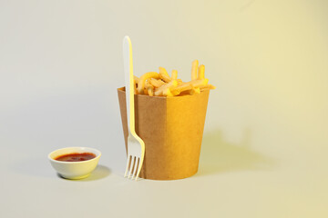Box with tasty french fries, fork and sauce on light background