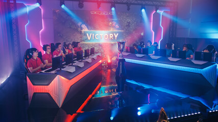 Two Esport Teams of Pro Gamers Play in RPG Strategy Video Game on a Championship Arena, Happy Red Team Wins Round and Celebrates with High-Fives. Big Screen Showing Victory Sign. Cyber Games Event © Gorodenkoff