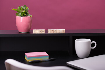 Close up view of a black work desk interior with a laptop computer, a cup of coffee,  paper, pen, plant and pink background.