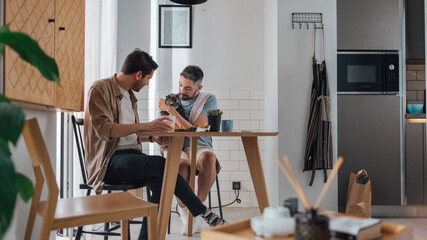 Happy Gay Couple Eat Healthy Breakfast Meal and Playing with Cat. Cheerful Young Boyfriends in Love...
