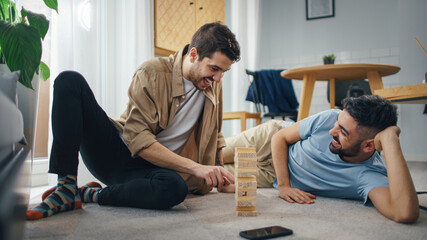 Happy Gay Couple Playing Wooden Block Tower Game while Resting on the Floor. Cheerful Young...