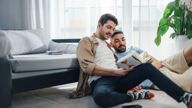 Happy Gay Couple Reading Book Together while Sitting on the Floor. Cheerful Young Boyfriends Enjoy Spending Time Together Having Fun, Talking. Authentically Tender Young Family Moment. - Powered by Adobe
