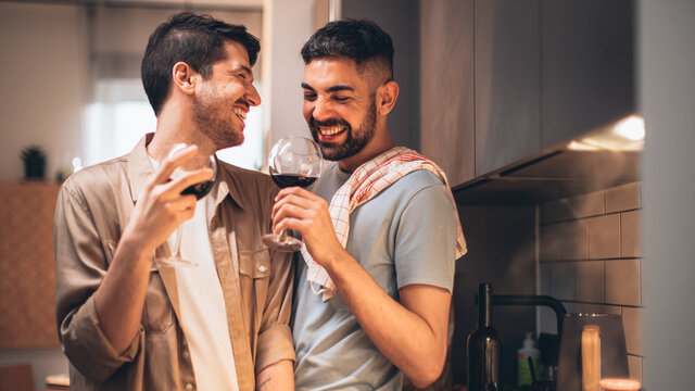 Happy Gay Couple In Love Drink Wine, Chat, Prepare Delicious Dinner Meal. Two Boyfriends Have Lovely Romantic Evening. Partners Talk, Share Beautiful Moments. Portrait Shot