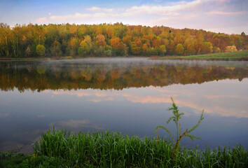Summer morning over the lake with misty haze on a background of forest and blue sky with clouds.
