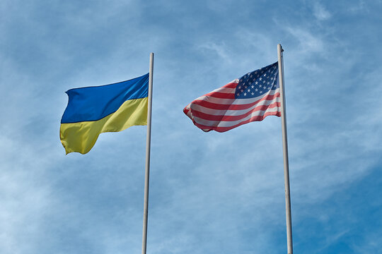 Flags of Ukraine and USA - United States of America waving in the wind at poles on cloudy sky background