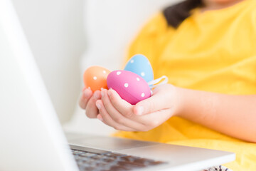 Happy easter day.Asian child girl in rabbit ears holding eggs.Online worship in easter holidays with laptop.Kid child online with bunny hat, easter egg hunt online at home.Lockdown, covid19, medical.