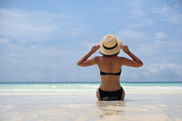 Fototapeta na wymiar Hot beautiful girl in a beach straw hat, hands raised to her head, black swimsuit sits on the beach on the beach with white sand and enjoys sunbathing under the gentle rays of the sun. Beach holidays