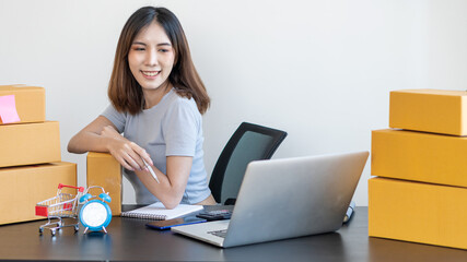 Young Asian businesswoman starts a small business, an online SME owner using a laptop, customizes customer orders with packaging boxes at home office and delivers their products online.