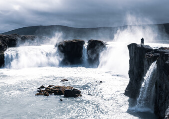 Obraz premium One man standing on a cliff facing the magnificent and tumultuous Goðafoss (waterfall of the gods) in a cool moody light. Iceland
