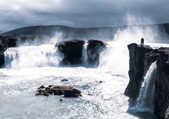One man standing on a cliff facing the magnificent and tumultuous Goðafoss (waterfall of the gods) in a cool moody light. Iceland