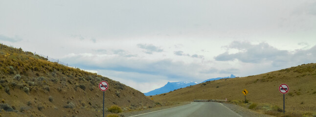 Photo of a road with traffic signs in a rural landscape with dry vegetation with snowy mountains in the background, cropped horizontal panoramic photo, double lane prohibited signs and curve sign