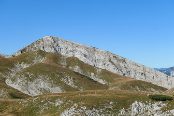 A panoramic view on Hochschwab mountain chains from the pathway leading to Hohe Weichsel. There is a vast pasture on top of a mountain, slowly turning golden. Clear view. Blue sky above. Autumn vibe