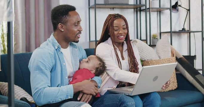 Handsome happy loving young black-skinned man holding his baby on hands sitting with his wife on sofa in contemporary living-room when another child running up to them
