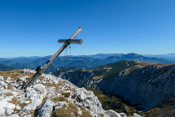 A wooden cross on top of Hohe Weichsel, Alpine peak in Austria. The cross is leaning, as if it was going to fall. There are endless mountain chains behind it. Early fall. The slopes are turning golden