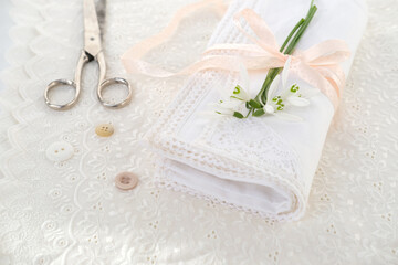 white linen napkin, trimmed with handmade lace, rolled in several layers, delicate snowdrop flower, scissors, buttons, ribbon is on light table, concept of needlework