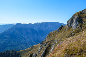 Obraz premium A panoramic view on Hochschwab mountain chains from the pathway leading to Hohe Weichsel. There is a vast pasture on top of a mountain, slowly turning golden. Clear view. Blue sky above. Autumn vibe
