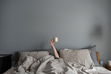 Woman laying in bed and holding mug with coffee with hand. Happy morning concept. Minimalist modern...