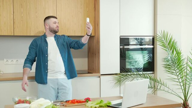 Happy young bearded man talking on the front camera of a mobile phone smartphone via video call standing in kitchen at home. Smiling handsome male waving hand hello into selfie camera meeting a family