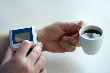 A man's hand with a blood pressure monitor on his wrist and a cup of coffee in his hand. Selective focus.