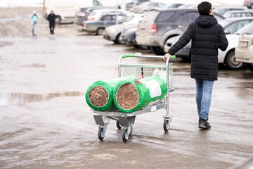  Parking near the construction shop. A young man pulls a cargo cart with thermal insulation material to the car.