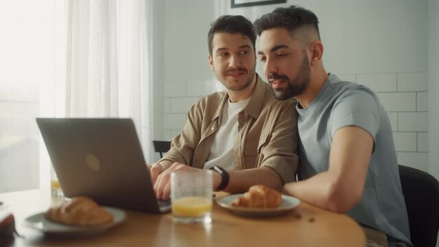 Happy Gay Couple Having Healthy Breakfast and Use Laptop. Sunday Morning Two Boyfriends in Love have Fun Talking, Watching Videos, Doing e-shopping. Partners Share Tender Moments
