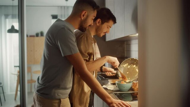 Happy Gay Couple Cooking Together in the Kitchen. Two Boyfriends in Love Spending Time Together. Boys Preparing Delicious Meal, Talk, Laugh and Have Fun. Authentically Tender Young Family Moment