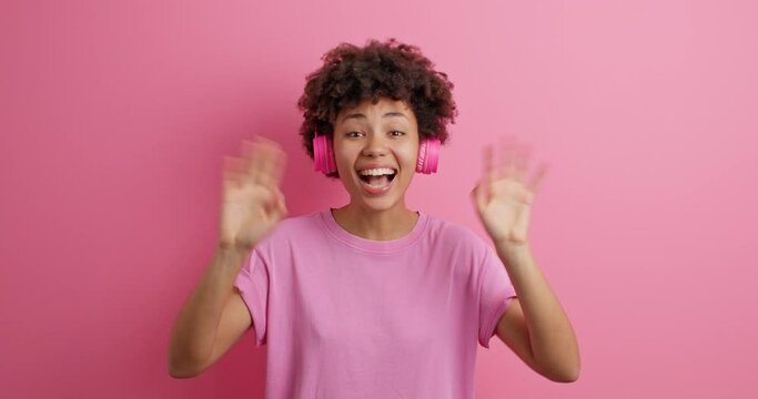 Beautiful cheerful Afro American woman waves hello smiles joyfully dressed in casual clothes enjoys listening to music dressed in casual clothes isolated over pink background. Hi to everybody
