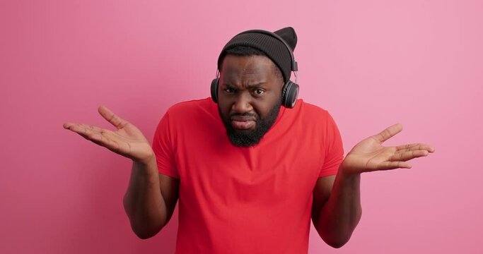 Displeased indignant bearded man spreads palms has no idea doesnt know what to do wears hat casual red t shirt isolated over pink background. Dissatisfied African American guy feels doubtful