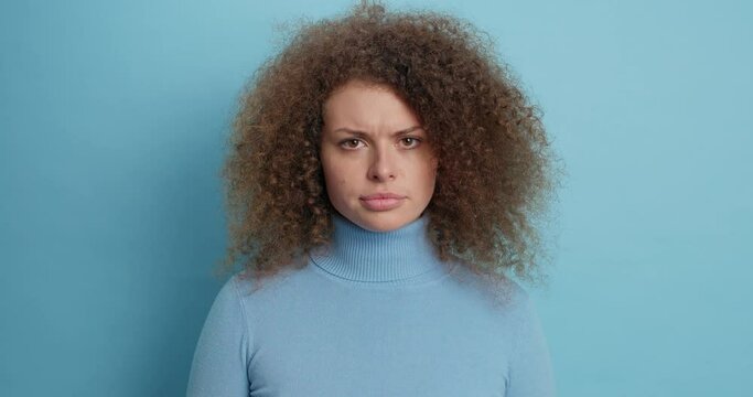 Dissatisfied curly haired European woman smirks face has sulking face expression raises eyebrows looks unhappy being discontent with something wears casual poloneck isolated over blue background