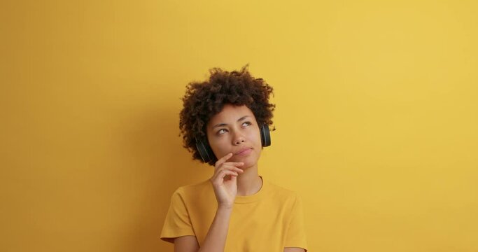Pensive dark skinned woman with curly hair looks around with thoughtful expression wears stereo headphones listens pleasant melody poses indoor against yellow backgroud. Daydreaming concept.
