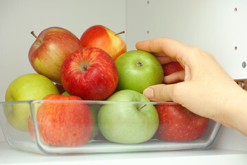 A woman taking a green apple out of a pile of apples that are in a glass tray that is in a kitchen cupboard