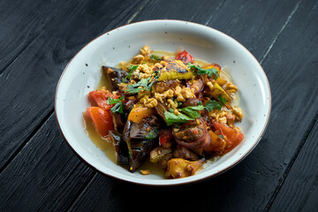 Appetizing, dietary salad of baked vegetables (tomatoes, peppers, eggplants) with sauce and nuts in a white plate on a dark wooden background. Vegetarian food. Selective focus