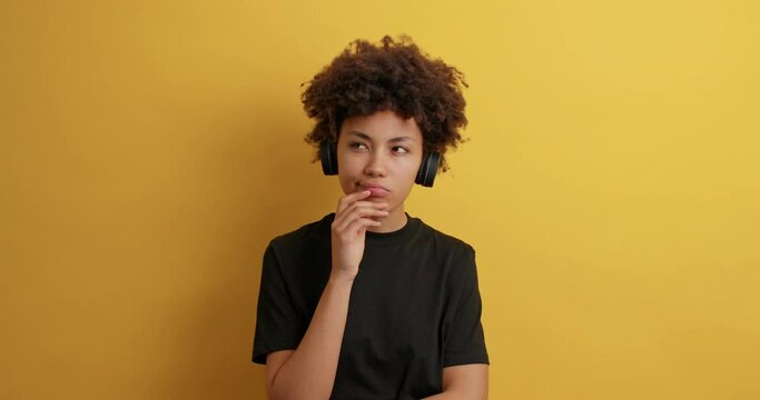Pensive curly haired woman looks around with thoughtful expression makes assumptions needs to think listens music via wireless headphones dressed in casual black t shirt isolated on yellow background