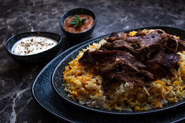 A plate of kabsa rice and grilled lamb and two small plates of Yogurt and hot sauce.