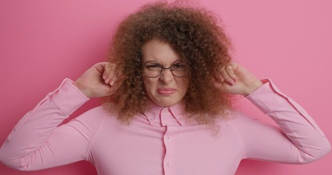 Annoyed curly haired woman asks to turn volume off irritated from bothering sound or loud music wears round spectacles and shirt isolated over pink background. Angry pissed off cant stand noise