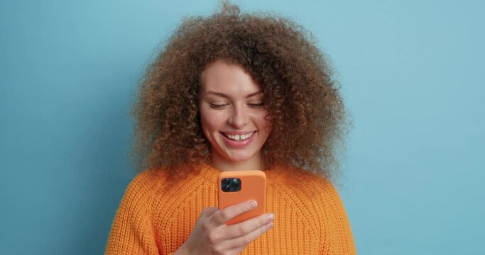 Smiling curly haired woman feels glad uses smartphone app texts friends or makes shopping online dressed in casual knitted sweater isolated over blue background. Communication technology concept